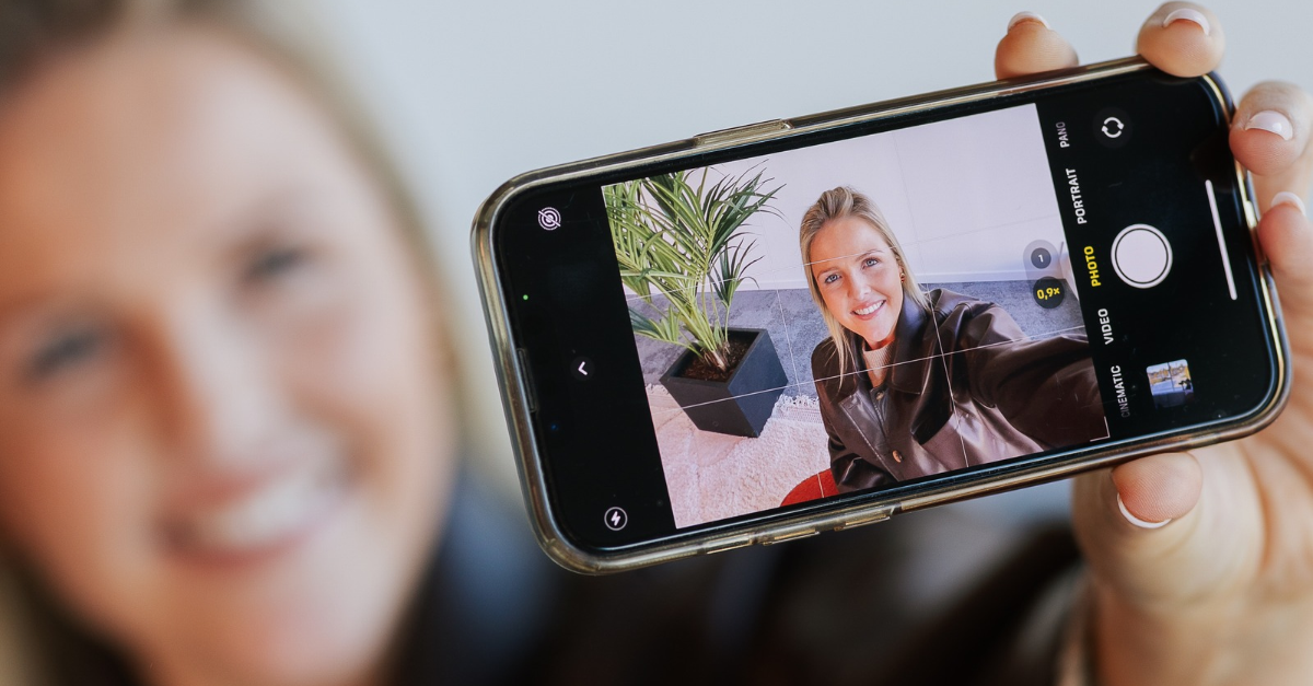 Glimlachende vrouw maakt een selfie met haar smartphone, waarbij haar gezicht scherp zichtbaar is op het telefoonscherm.