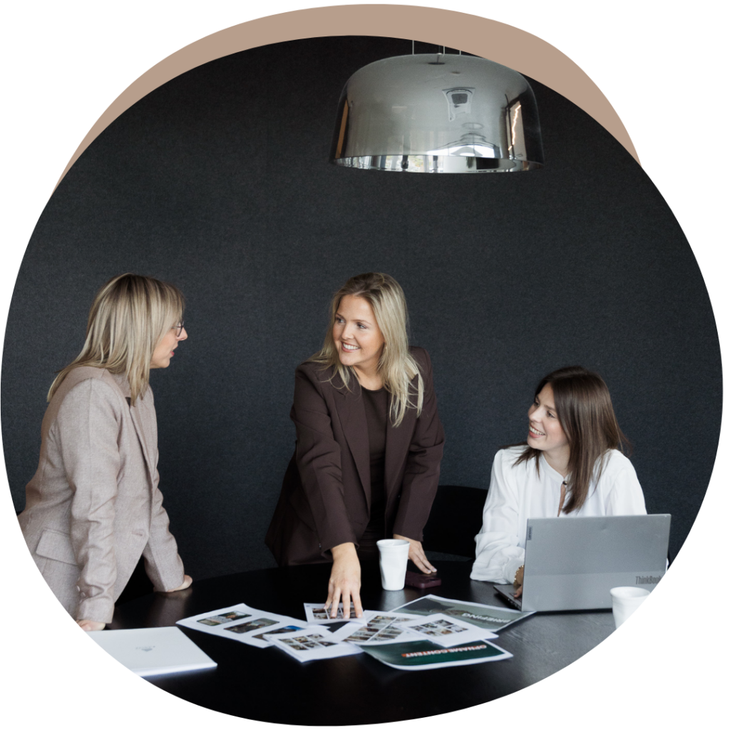 Drie vrouwelijke collega's van E-Volve overleggen aan een vergadertafel met een laptop en documenten over marketing en strategie.