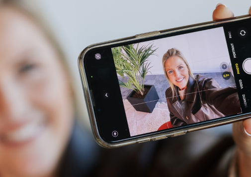 Glimlachende vrouw maakt een selfie met haar smartphone, waarbij haar gezicht scherp zichtbaar is op het telefoonscherm.