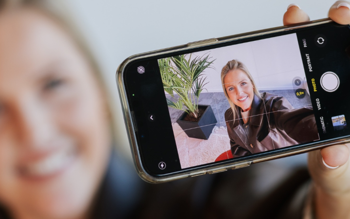 Glimlachende vrouw maakt een selfie met haar smartphone, waarbij haar gezicht scherp zichtbaar is op het telefoonscherm.